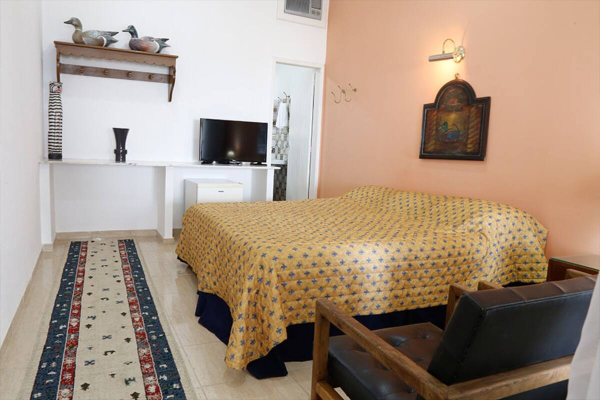 Quarto com uma cama, televisão, prateleira com objetos decorativos, luminária na parede e uma porta.