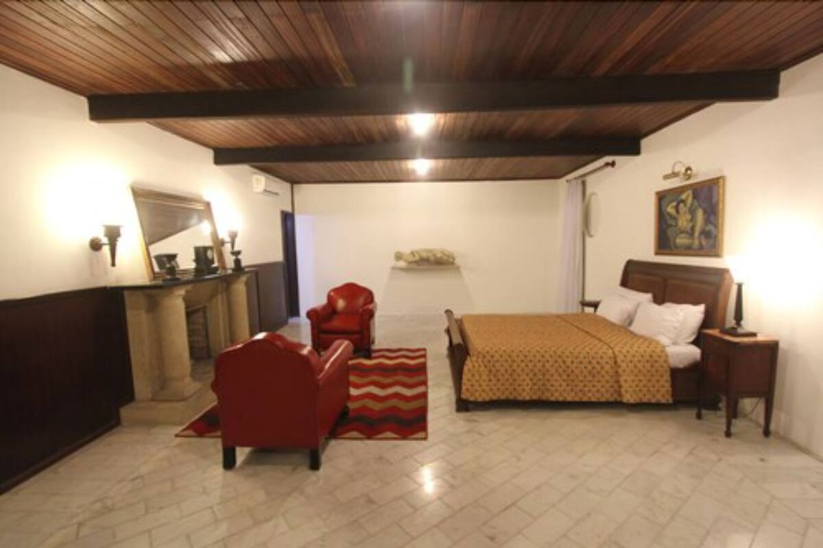 Quarto com cama, poltrona vermelha, teto de madeira e um quadro na parede.