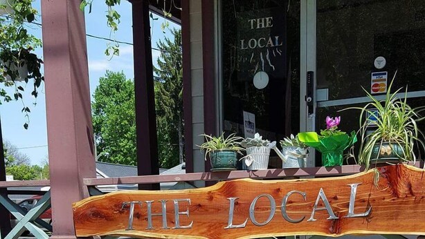 Une enseigne en bois sur laquelle on peut lire « THE LOCAL », placée au-dessus d’un banc devant un bâtiment dont le mur est en briques.