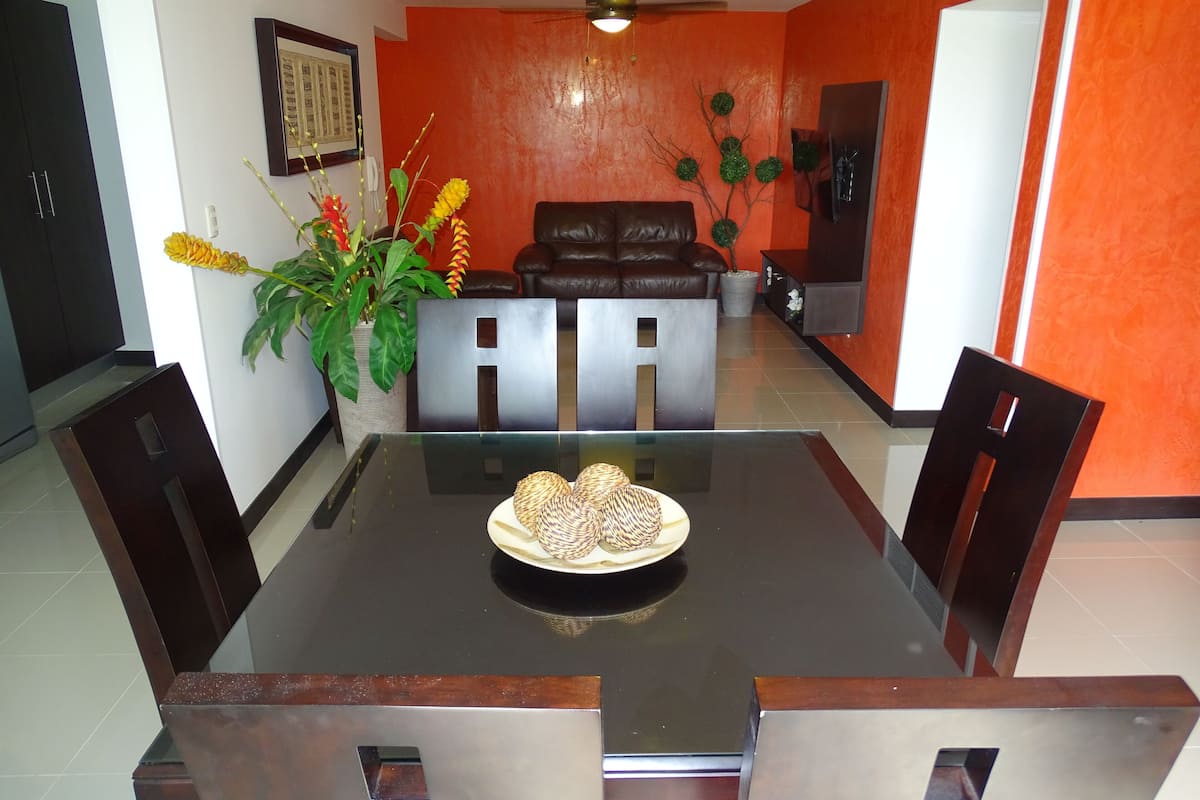 Un comedor con mesa de vidrio, cuatro sillas, un sofá y una planta decorativa.