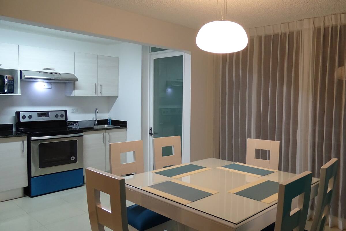 Una cocina compacta con estufa, horno y microondas, una mesa de comedor con sillas y una ventana con cortina.