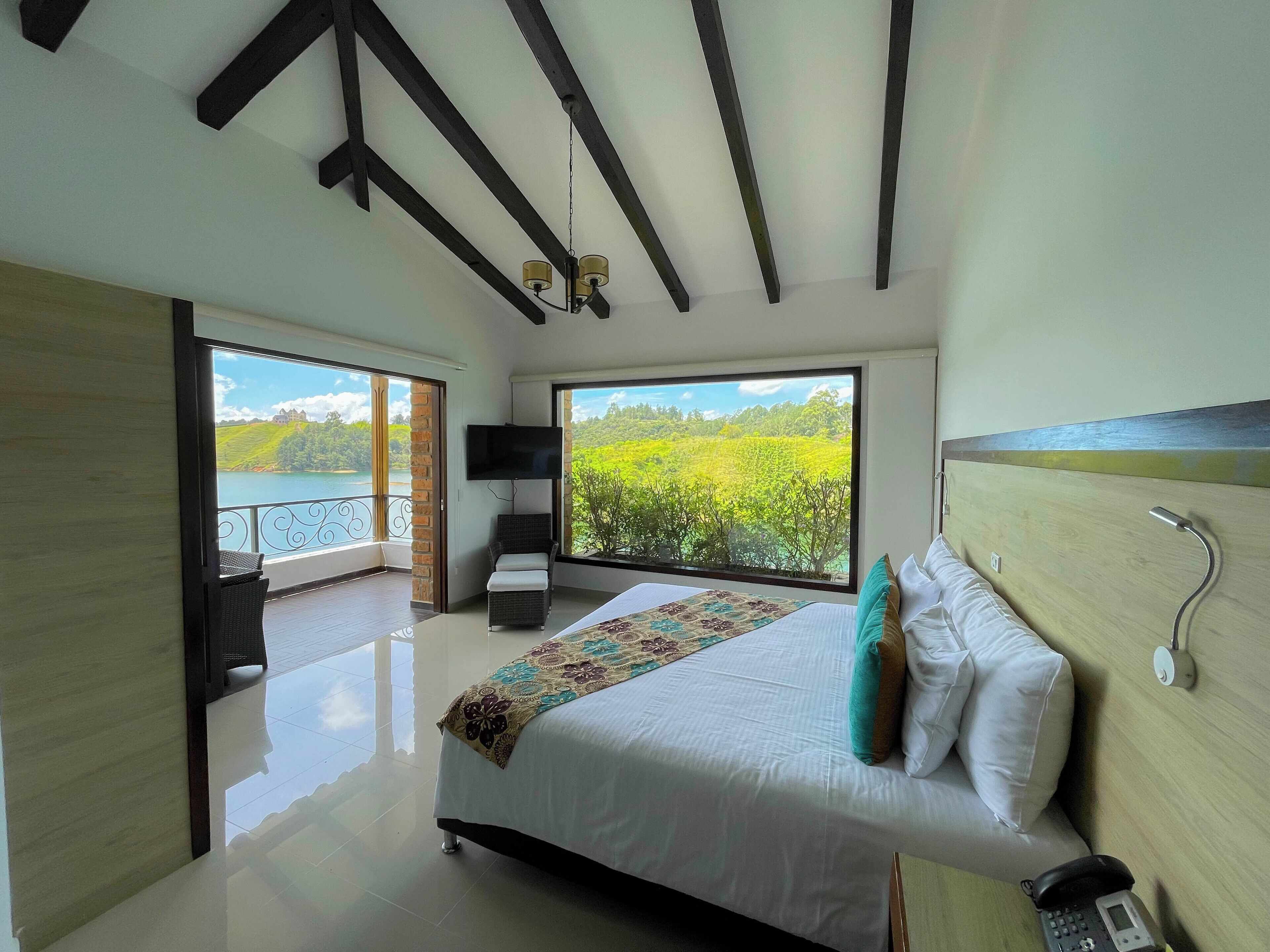 A bedroom with a large bed, a TV, a bedside table, and a view of a lush landscape.