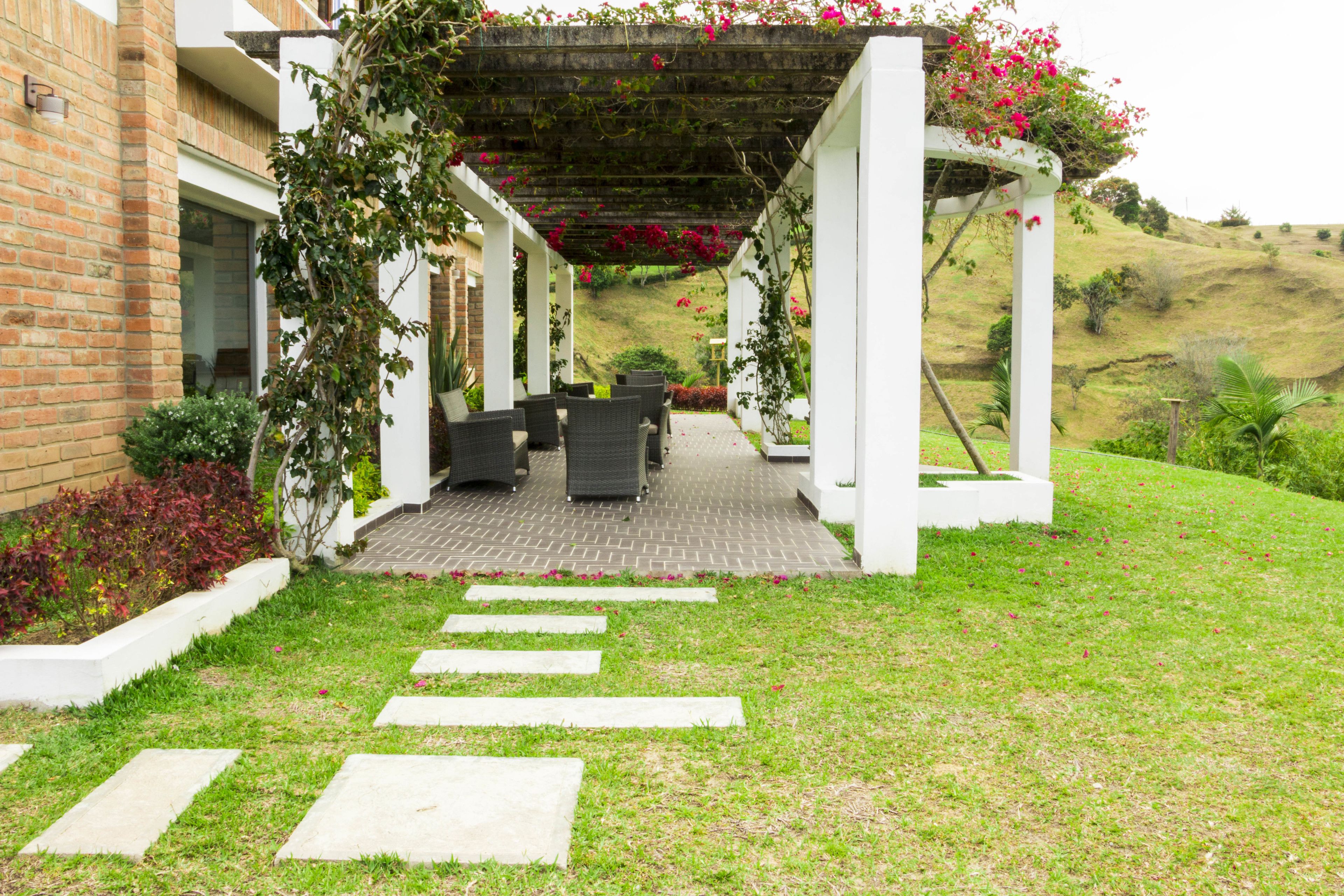 A covered outdoor seating area with stone steps leading to it, surrounded by greenery and a hillside in the background.