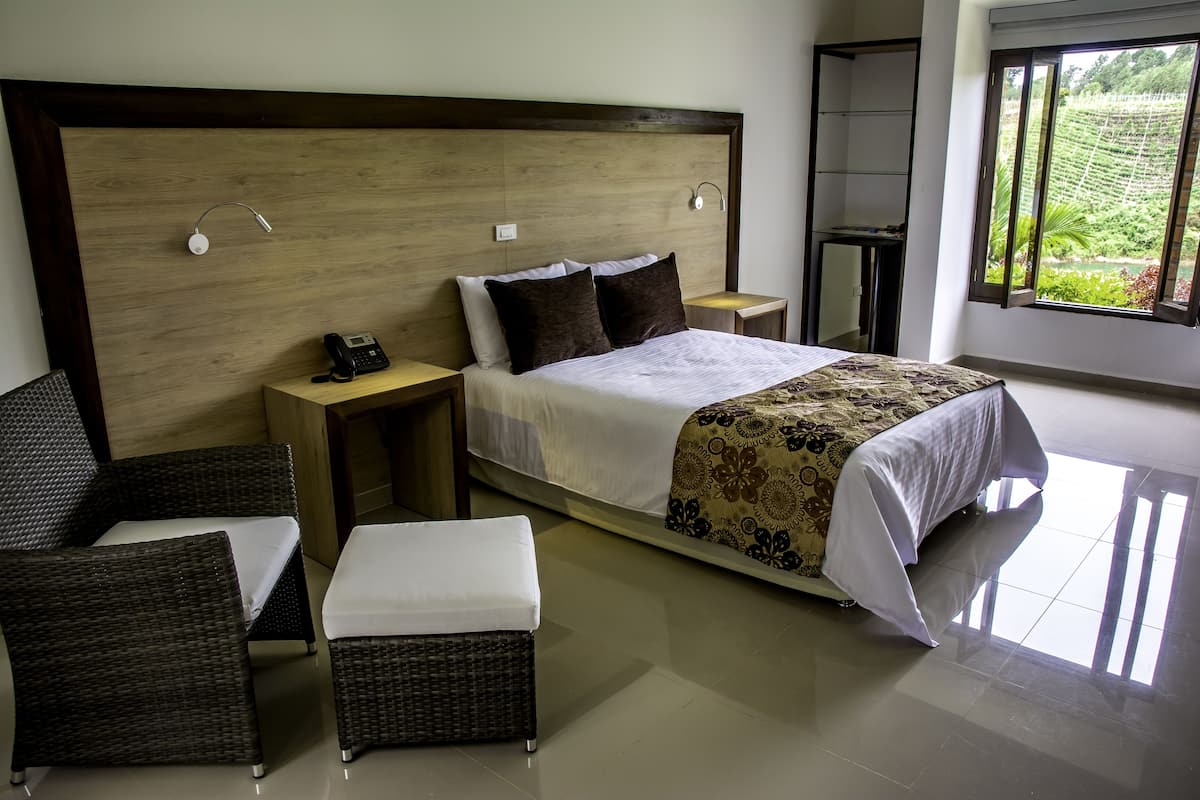 Ein Schlafzimmer mit einem hölzernen Kopfteil, einem Bett mit weißer Bettwäsche, zwei Nachttischen, einem Telefon, einem kleinen Tisch, zwei Korbstühlen und einem großen Fenster mit Blick auf Grünflächen.