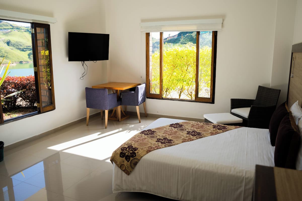 Ein Hotelzimmer mit Bett, Schreibtisch, Stuhl, Fernseher und großen Fenstern mit Blick auf Grünflächen.