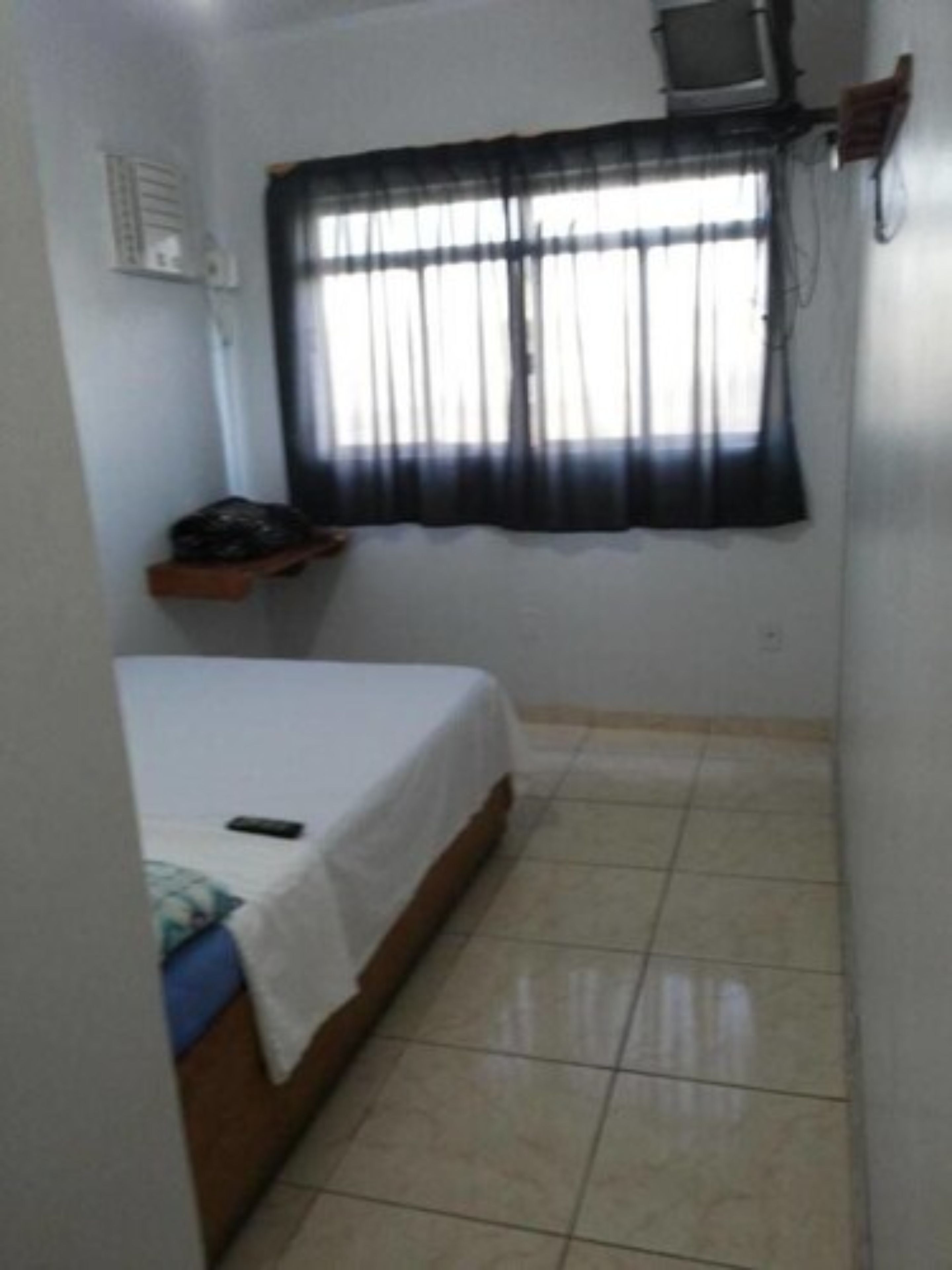 Quarto pequeno e limpo, com cama, janela com cortinas leves, prateleira pequena e ar-condicionado fixado na parede.