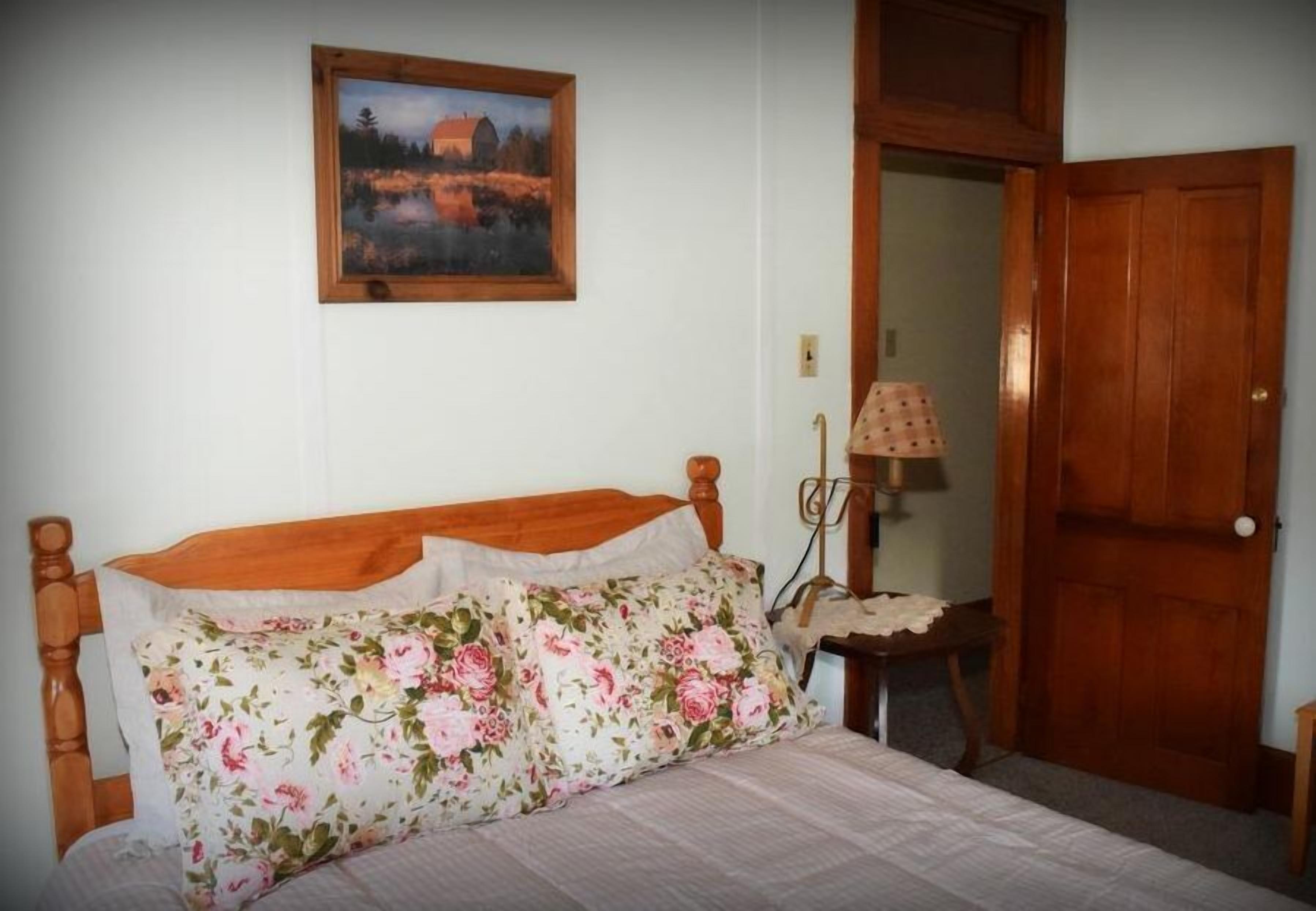 A bedroom with a wooden bed, floral bedding, a painting on the wall, a bedside lamp, and a wooden door.