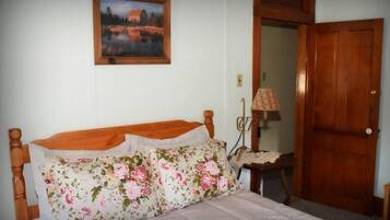 A bedroom with a wooden bed, floral bedding, a painting on the wall, a bedside lamp, and a wooden door.