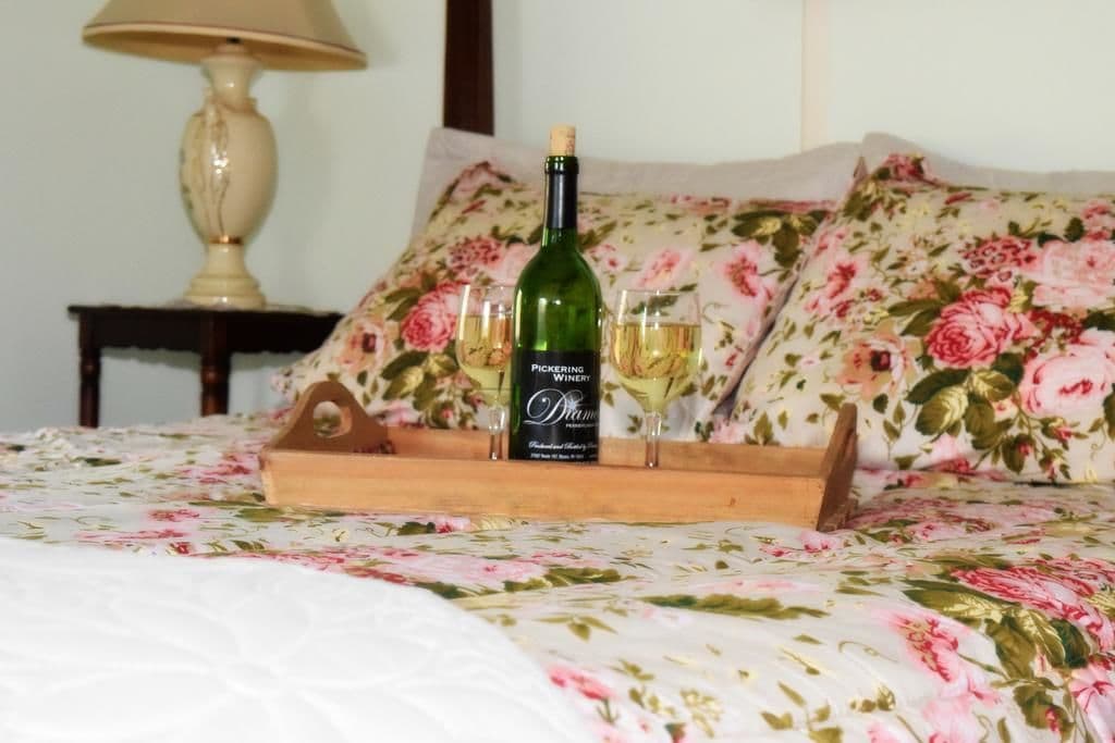 A bed with a floral bedspread, a bottle of Pickering Dream wine, and two glasses on a wooden tray.