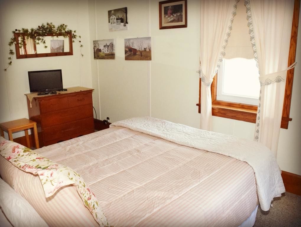 Un dormitorio con una cama grande, un aparador de madera, un televisor sobre un mueble bajo y una ventana con cortinas.
