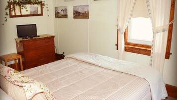 Un dormitorio con una cama grande, un aparador de madera, un televisor sobre un mueble bajo y una ventana con cortinas.