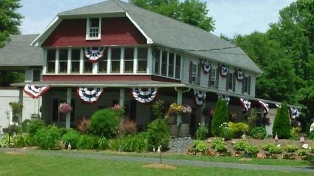 A two-story building with a red facade and a grey roof, featuring a porch with American flags and flower baskets. - Beagle's Bed & Breakfast (Paxinos)