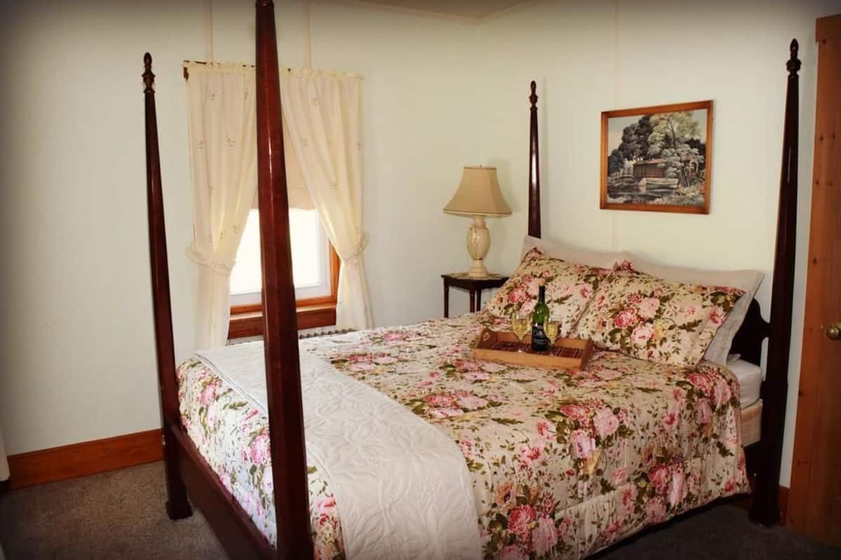 Un lit à baldaquin avec une literie à motifs floraux, une table de chevet avec une lampe et un tableau représentant un paysage, encadré, accroché au mur.