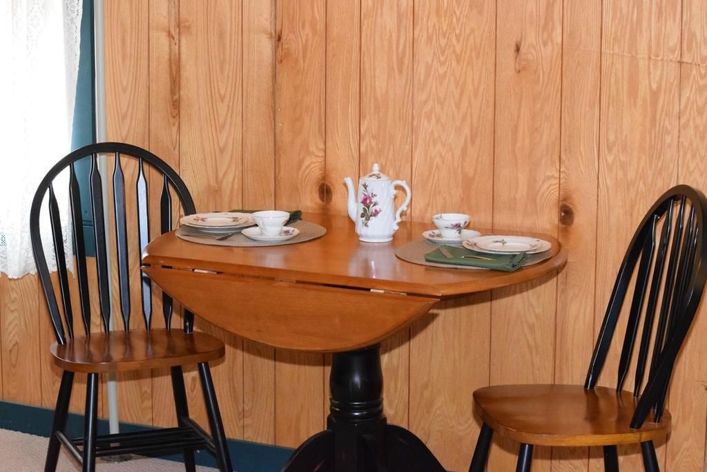A wooden table with a white teapot and matching cups, flanked by two black chairs.