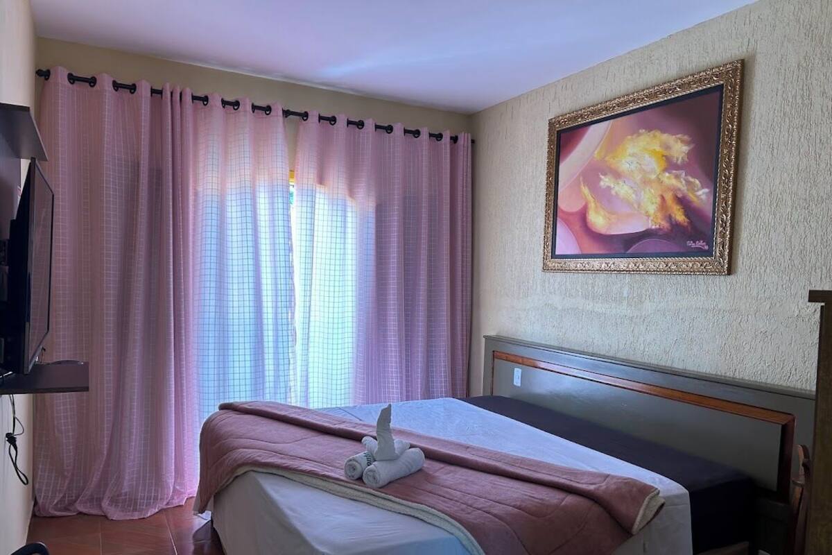 A hotel room with a bed, a television, a painting on the wall, and a window with curtains.
