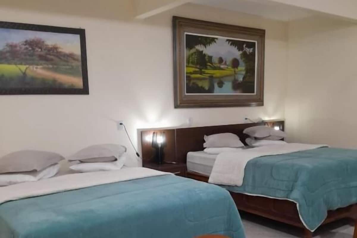 A hotel room with two beds, a nightstand, a lamp, two wooden stools, and two paintings on the walls.