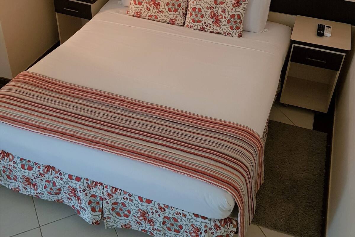 A neatly made bed with floral-patterned bedding, a bedside table with a remote control, and a framed picture on the wall.