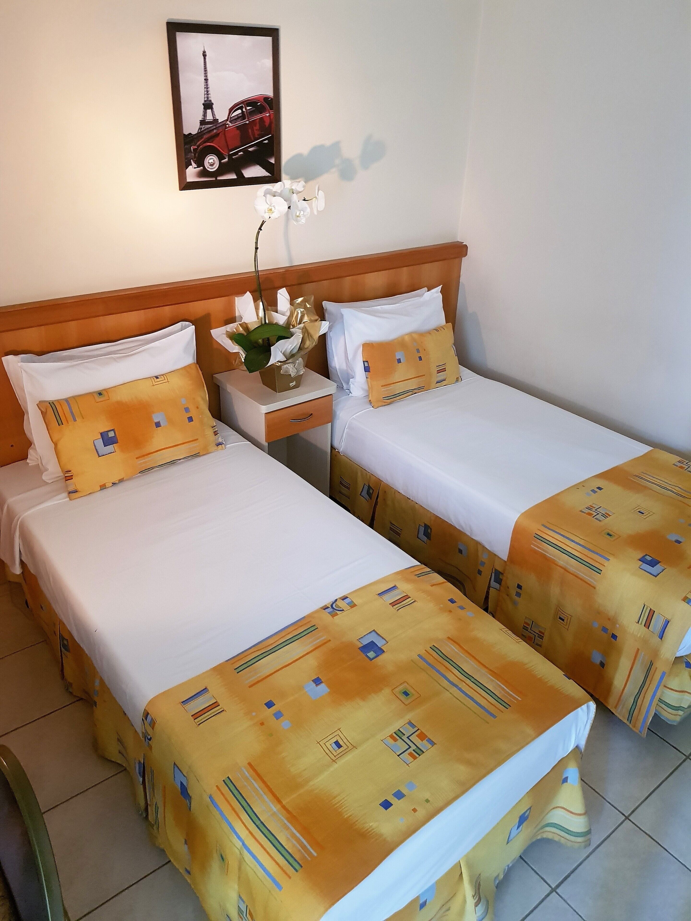 Quarto de hotel com duas camas, uma cabeceira de madeira, mesa de cabeceira com um vaso de flores e uma moldura com a Torre Eiffel.
