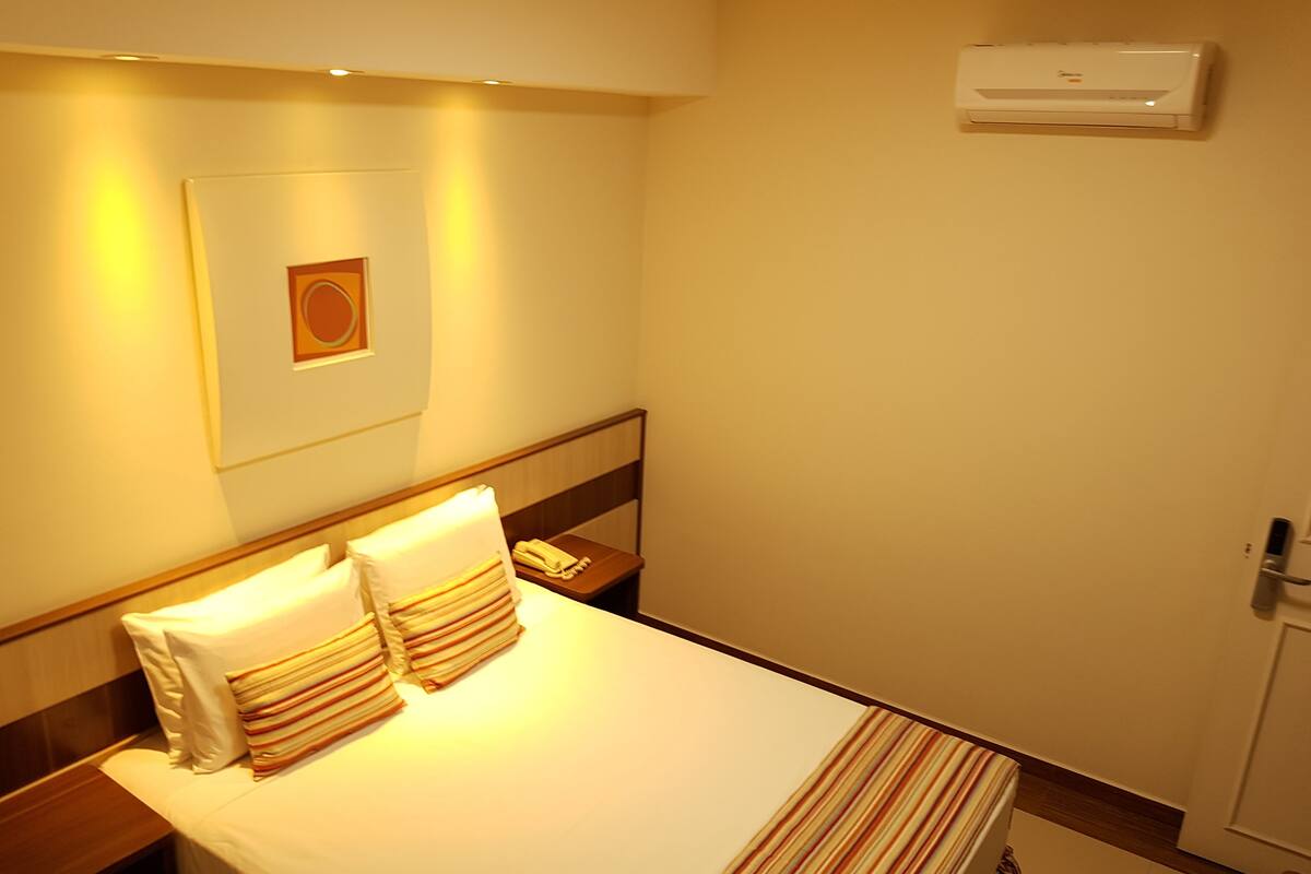 A hotel room with a bed, a bedside table, a telephone, an air conditioning unit, and a door.