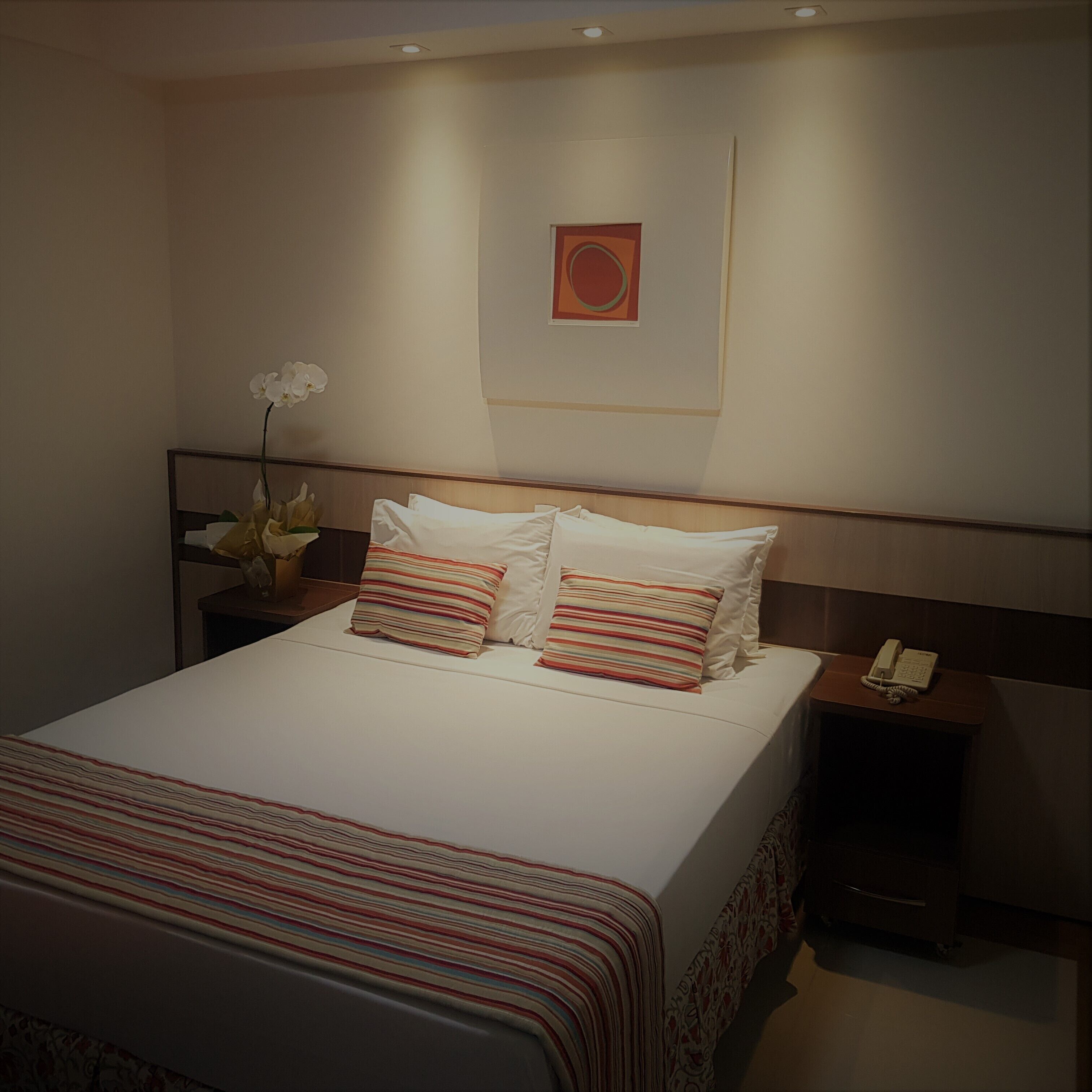 A neatly made bed with striped bedding, a bedside table with a telephone, and a wall-mounted artwork.