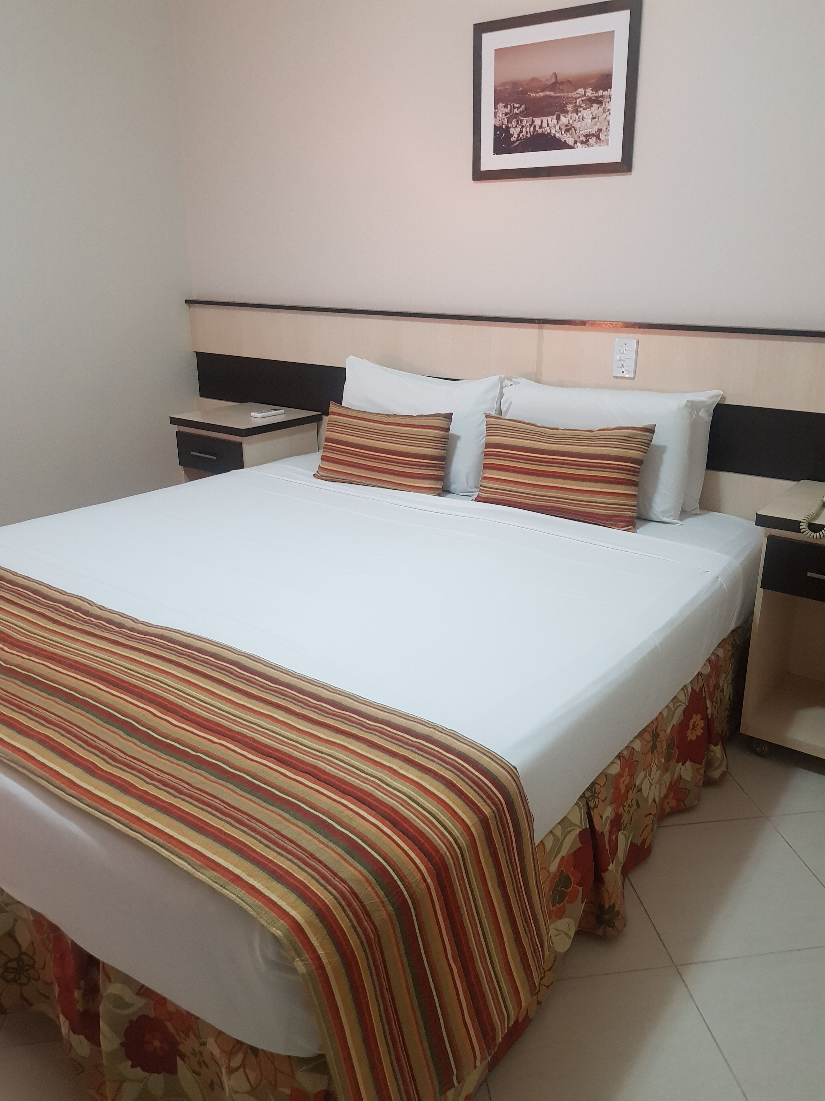 A hotel room with a bed, a nightstand, a framed picture on the wall, and a floral-patterned skirt on the bed.