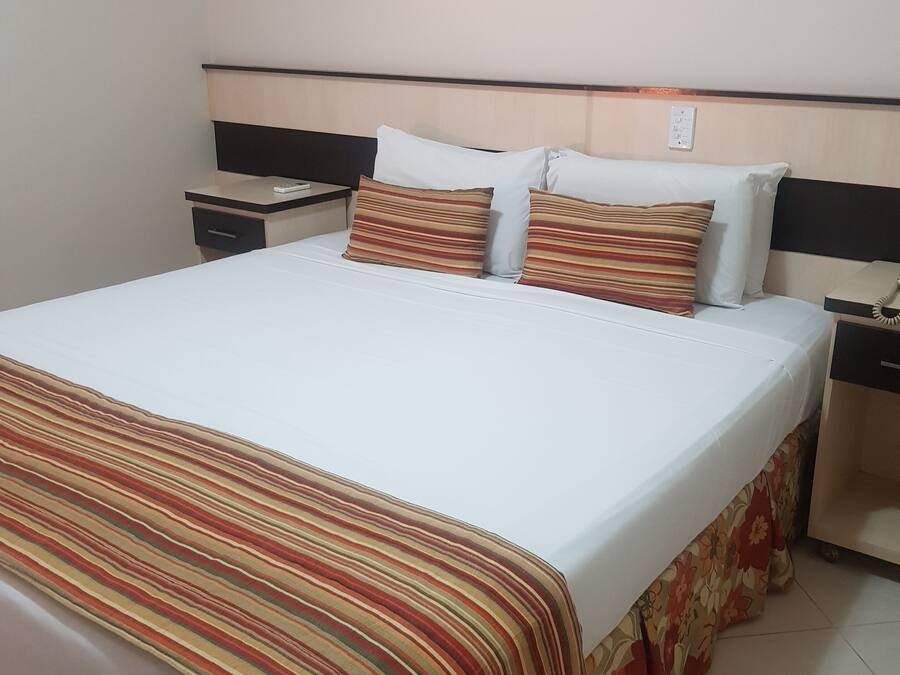 A hotel room with a bed, a nightstand, a framed picture on the wall, and a floral-patterned skirt on the bed.