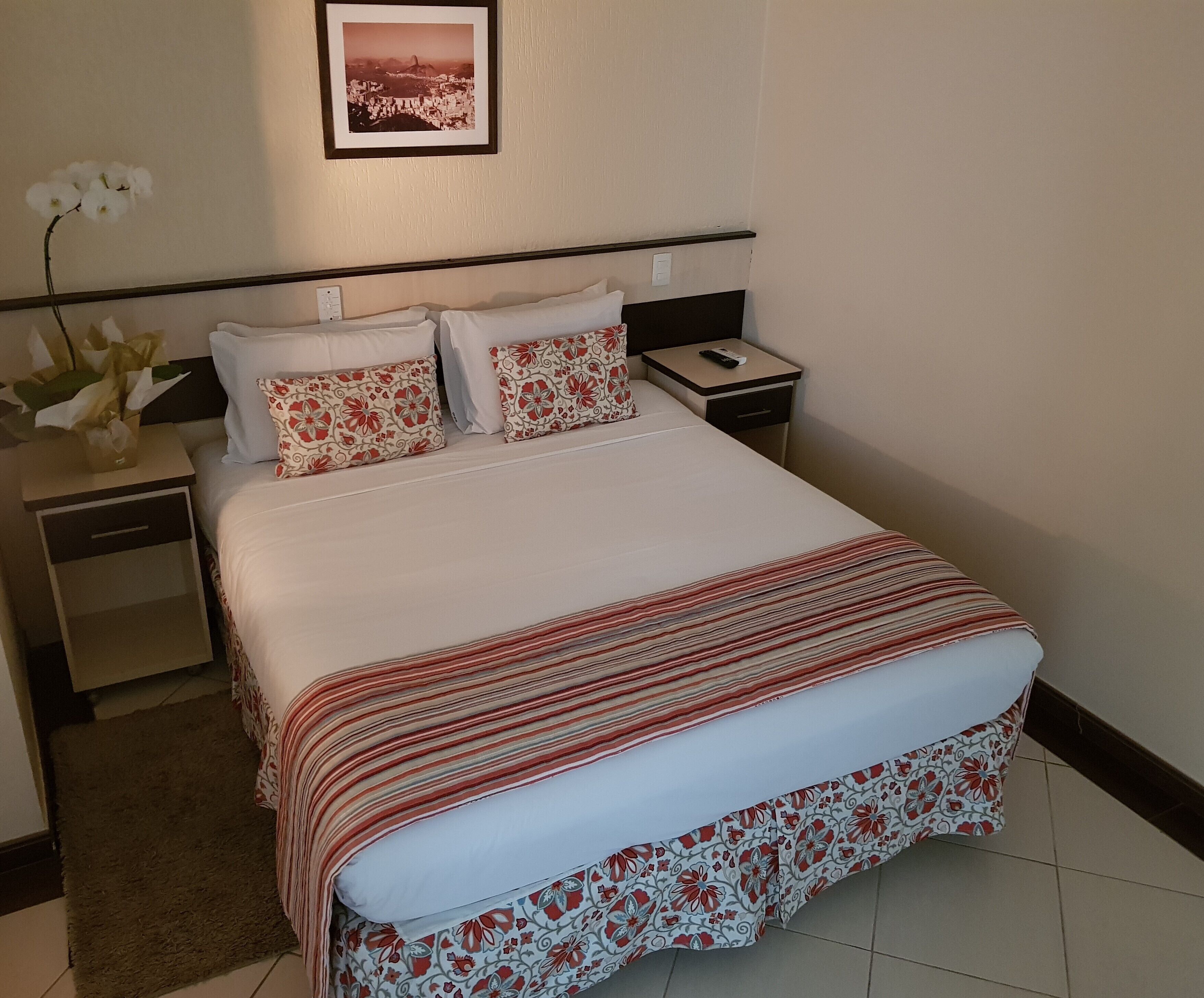 A neatly made bed with a striped bedspread, two pillows with a floral pattern, a nightstand with a remote control, and a framed picture on the wall.
