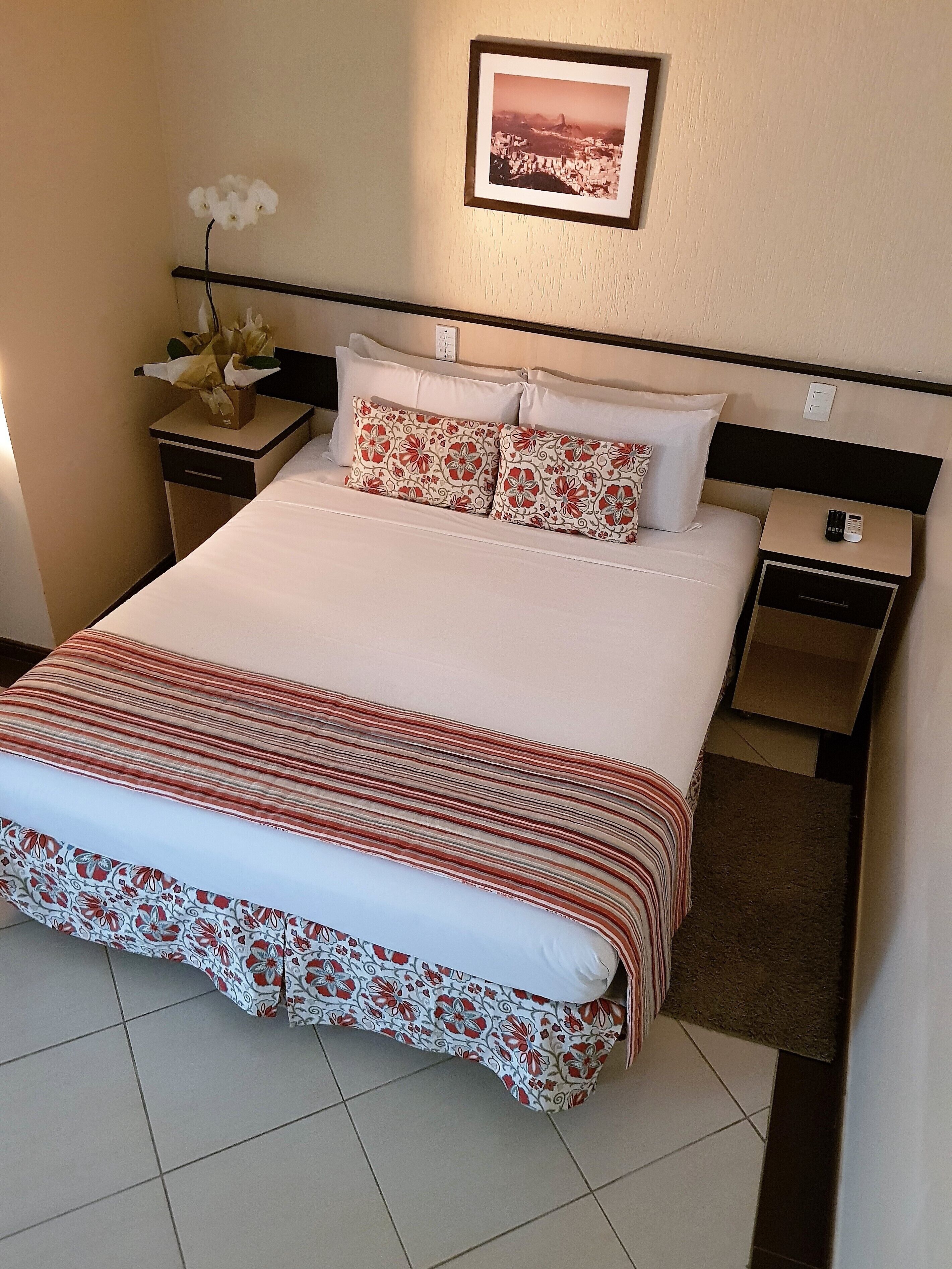 Una cama bien tendida con ropa de cama estampada con motivos florales, una mesita de noche con un control remoto y un cuadro en la pared.