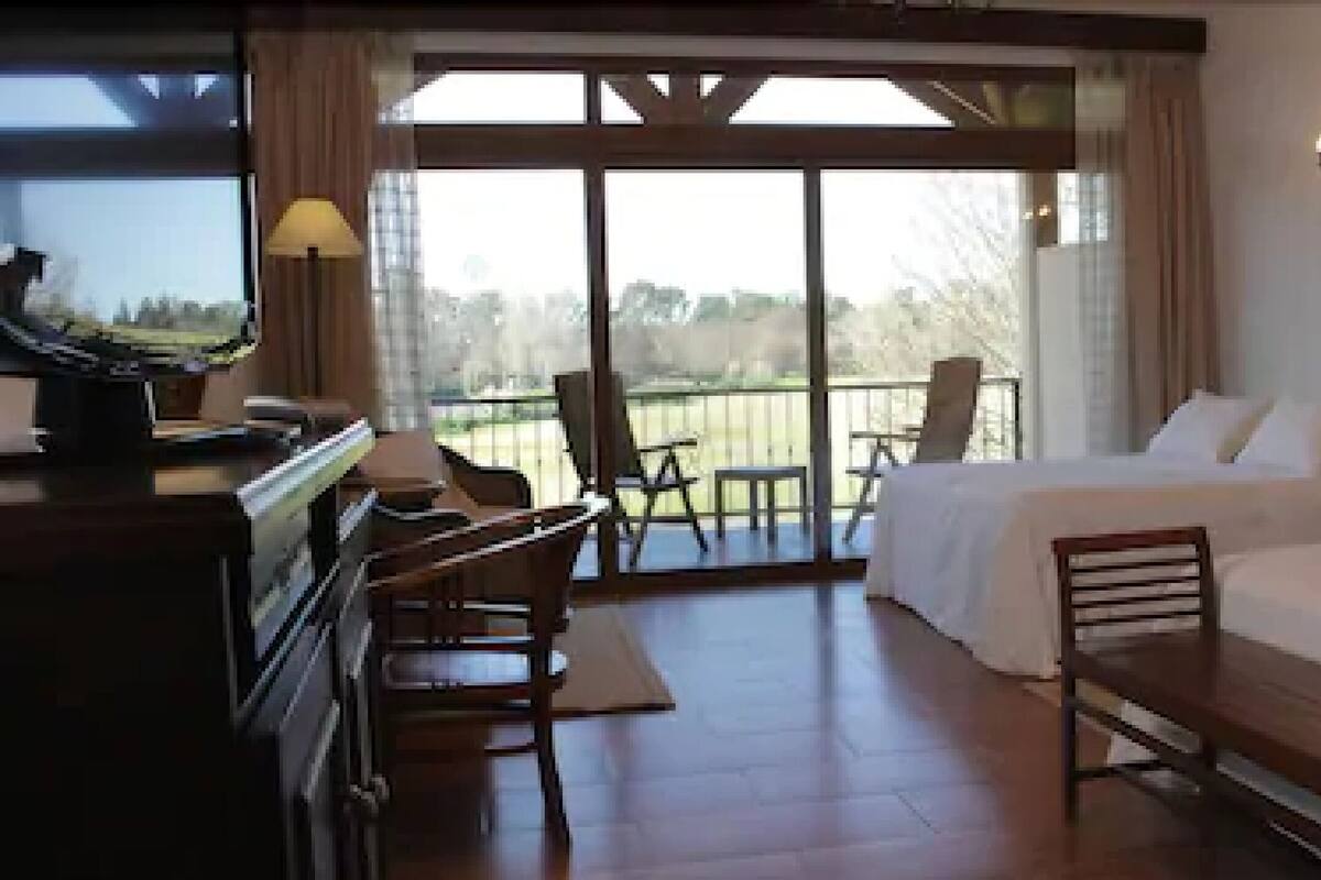 A hotel room with two beds, a desk, a piano, and a balcony with chairs.
