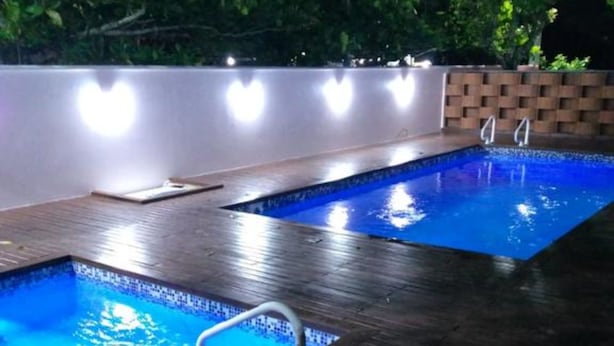 Una piscina exterior de noche con una cubierta de madera y un muro de ladrillo al fondo.