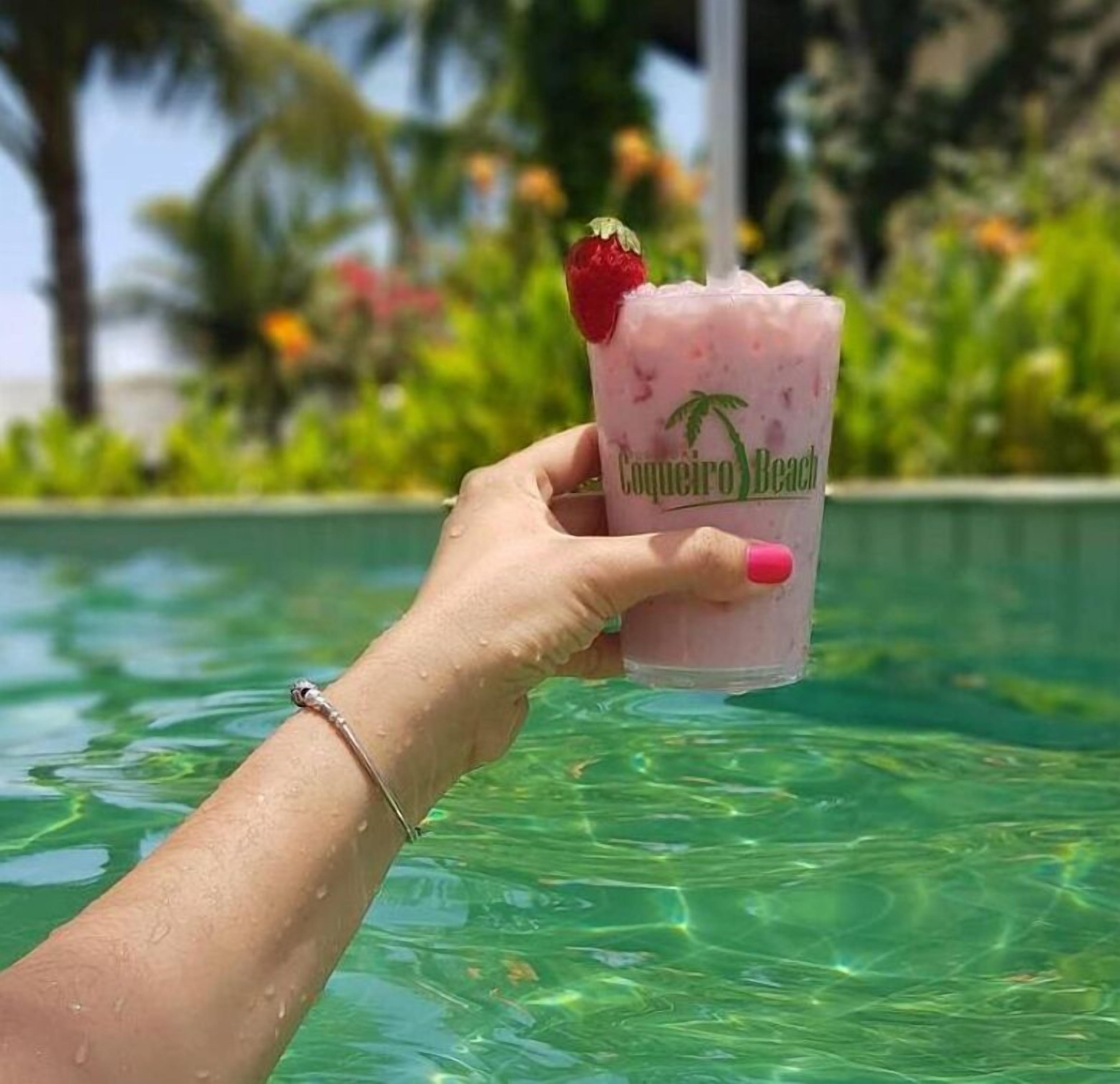 A hand holding a drink with a straw and a strawberry garnish, with a pool and greenery in the background.