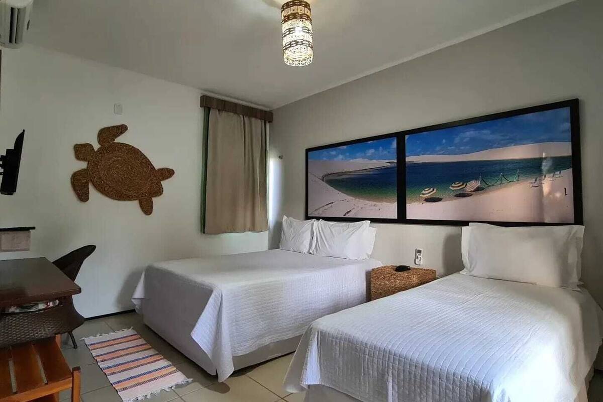 A hotel room with two beds, a desk, a turtle wall decoration, a beach view painting, and a ceiling light.