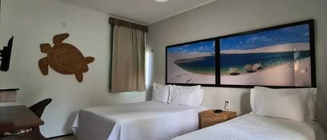 A hotel room with two beds, a desk, a turtle wall decoration, a beach view painting, and a ceiling light.