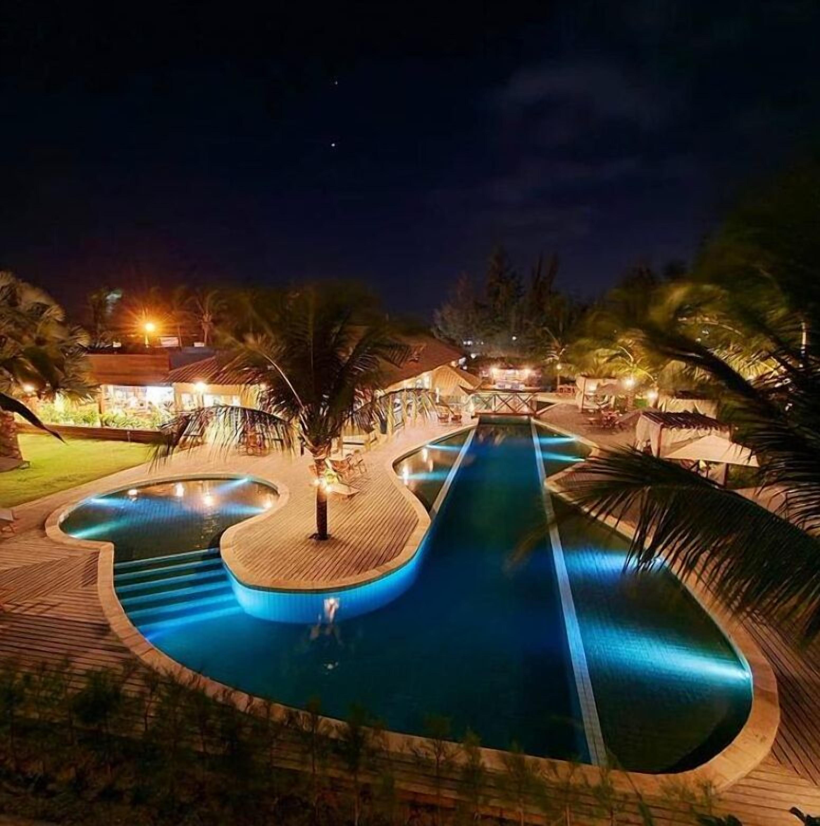 A nighttime view of a resort with a central swimming pool, palm trees, and illuminated buildings.