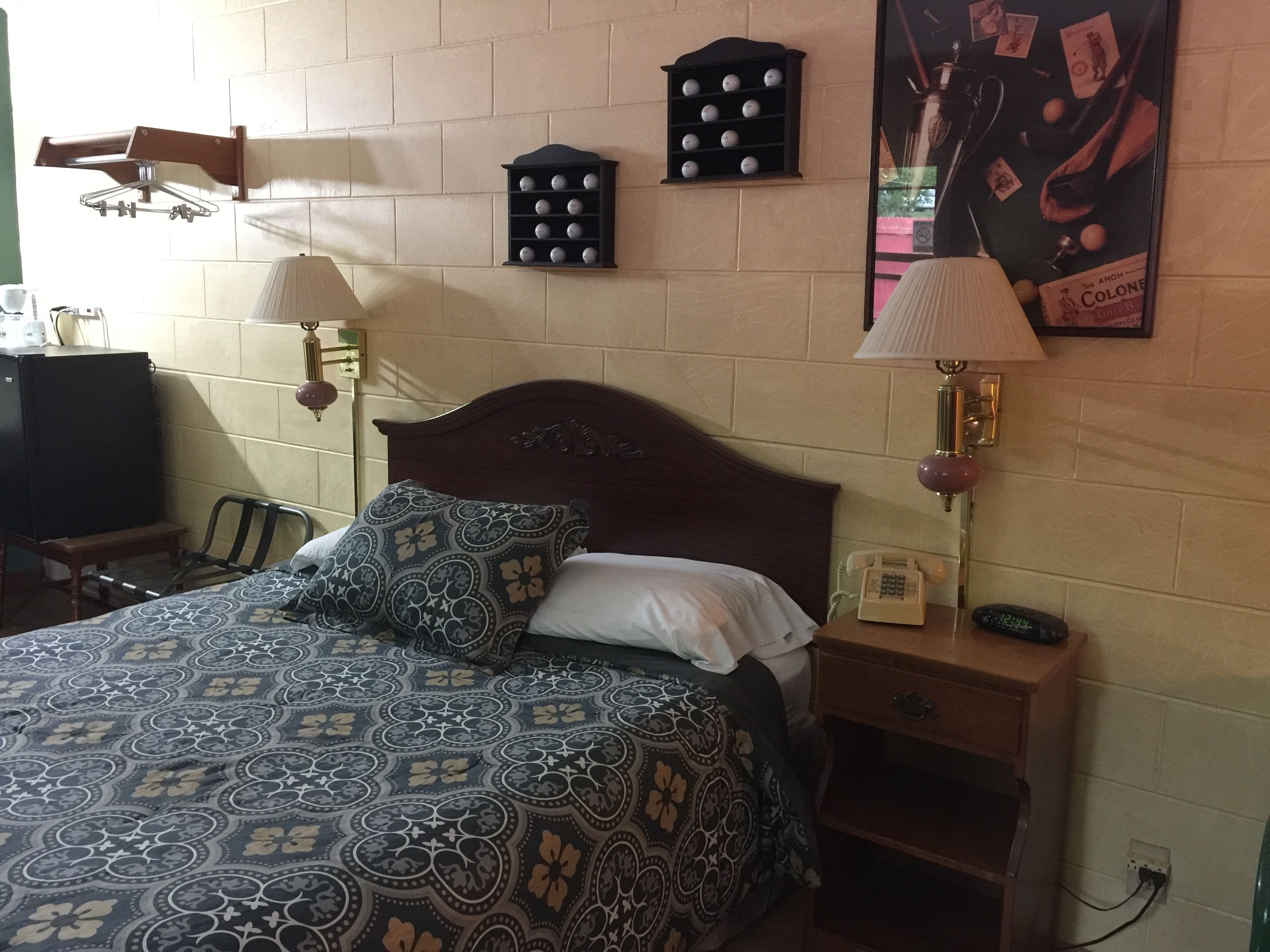 A neatly made bed with patterned bedding, a bedside table with a lamp, a small shelf with decorative items, and a wall-mounted telephone.