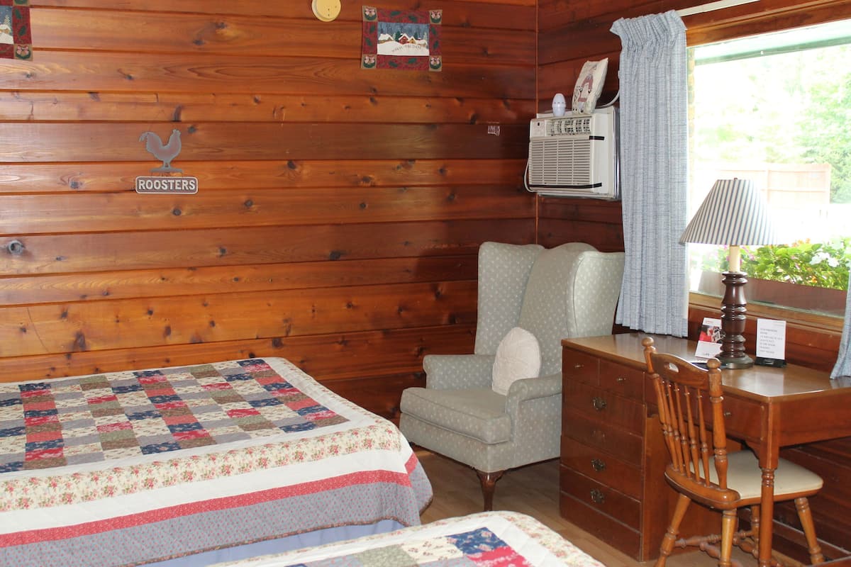 A cozy cabin room with a wooden wall, a quilted bed, a chair, a desk, and a window with curtains.