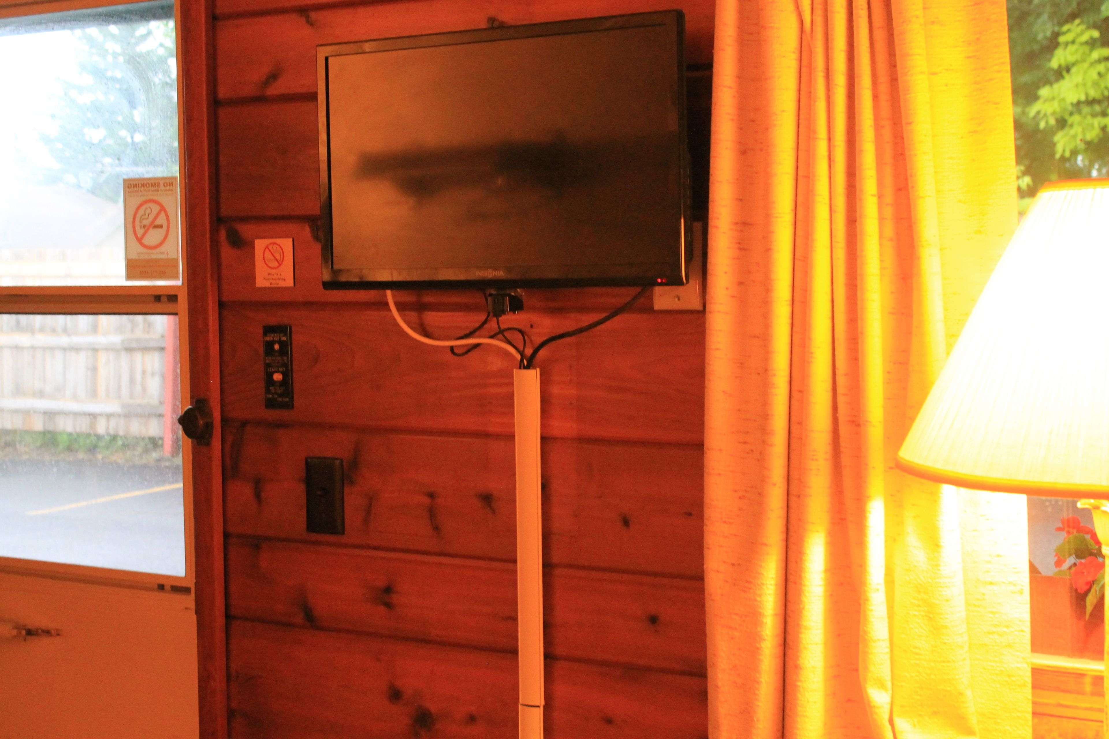 Un téléviseur fixé à un mur en bois, une fenêtre donnant sur une rue et une lampe sur la droite.