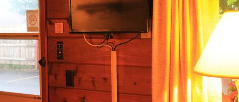Un téléviseur fixé à un mur en bois, une fenêtre donnant sur une rue et une lampe sur la droite.
