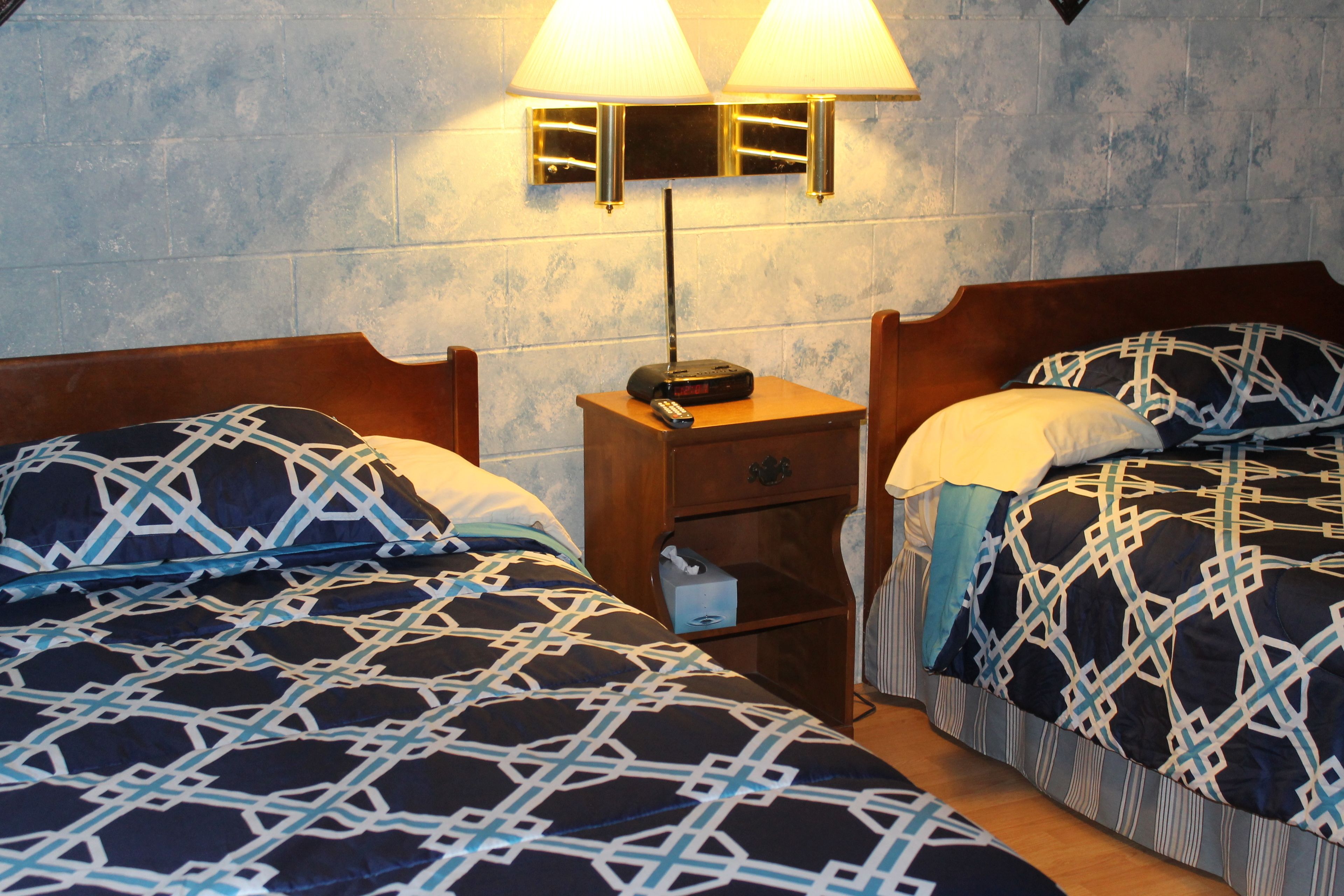 Two beds with patterned bedding, a nightstand with a lamp, and a small drawer unit.
