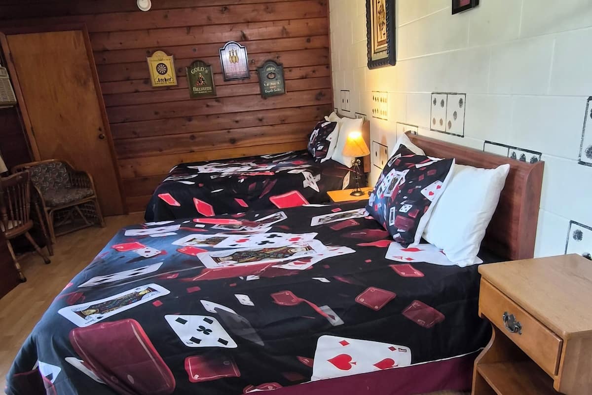 A bedroom with a wooden floor, a bed with a card game themed bedspread, a nightstand, and a chair.