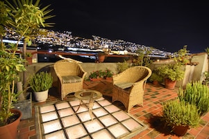 Terraza en la azotea con mobiliario de mimbre, plantas en macetas y vistas a la ciudad de noche.