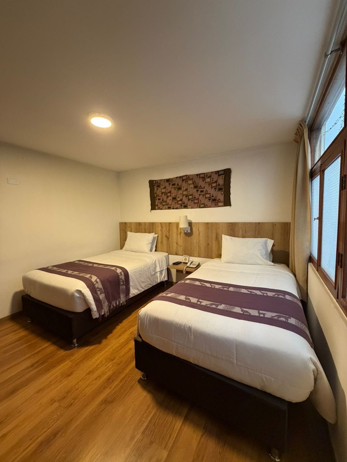 A hotel room with two beds, a wooden headboard, a wall-mounted telephone, and a window with curtains.