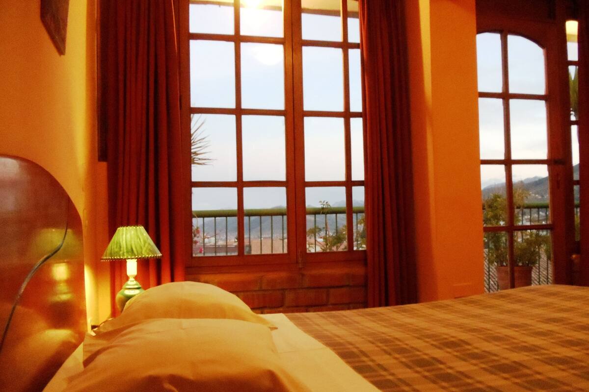 A hotel room with a large window, a bed, and a nightstand with a lamp.
