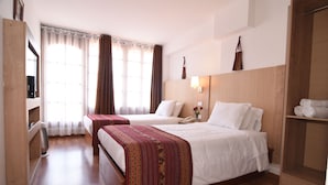 Une chambre d’hôtel avec deux lits, une armoire en bois, une table de chevet avec une lampe et une grande fenêtre avec des rideaux.