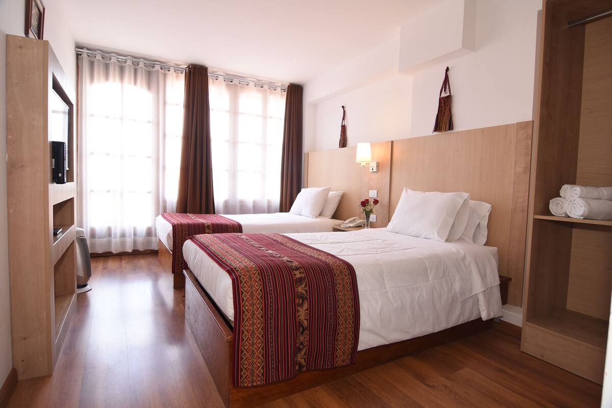 A hotel room with two beds, a wooden wardrobe, a bedside table with a lamp, and a large window with curtains.