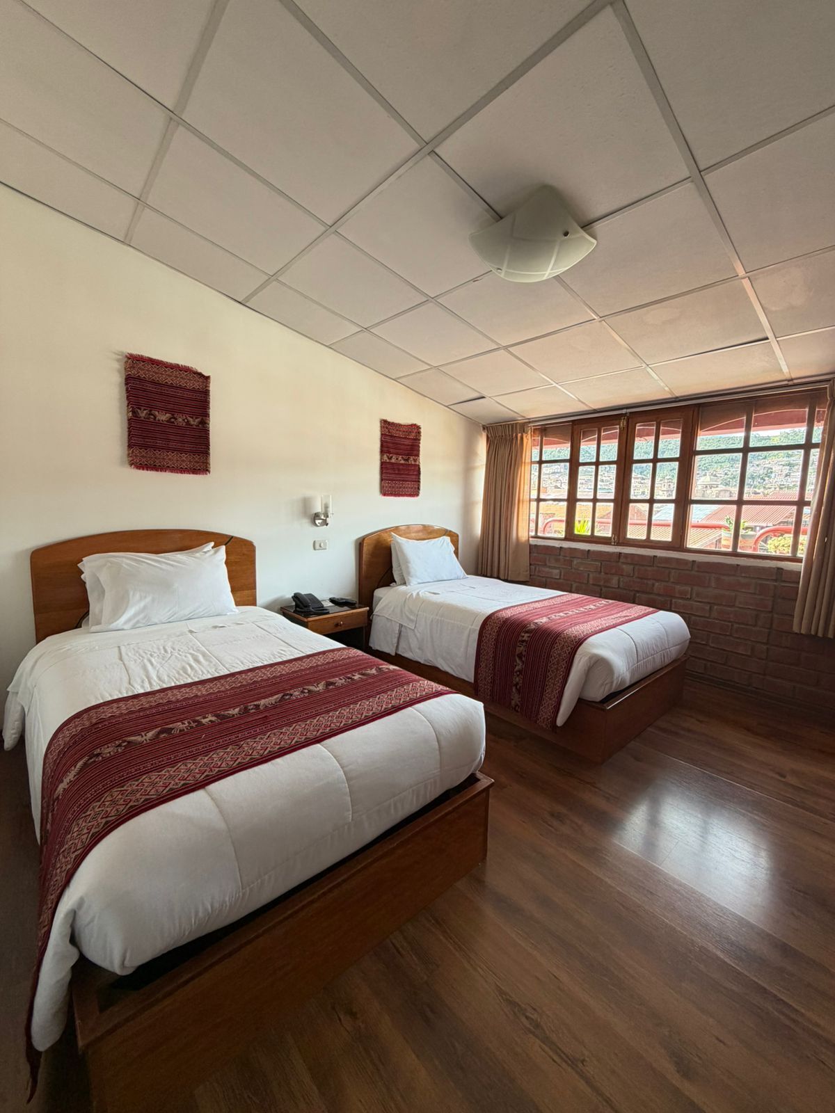 A hotel room with two beds, a wooden headboard, a nightstand, a telephone, a wall-mounted light, and a window with curtains.