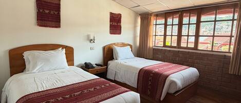 A hotel room with two beds, a wooden headboard, a nightstand, a telephone, a wall-mounted light, and a window with curtains.