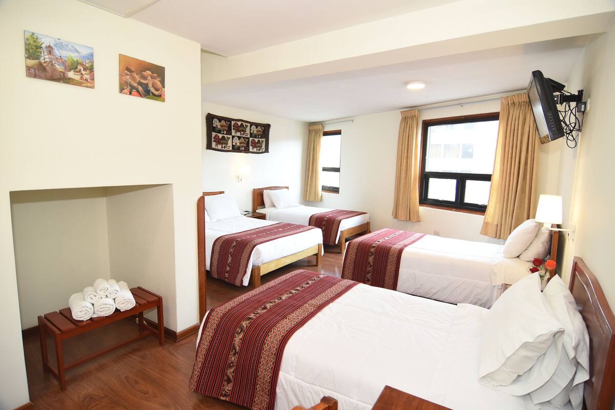 A hotel room with two beds, a bench with towels, a TV mounted on the wall, and artwork on the walls.