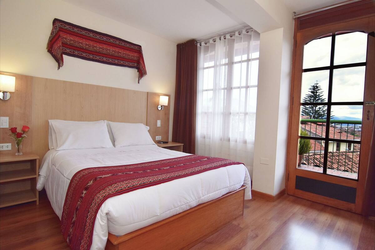 A bedroom with a wooden bed, white bedding, a red patterned throw, two bedside lamps, a nightstand, a window with curtains, and a view of greenery and buildings outside.