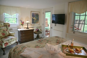 A bedroom with a floral bedspread, a dresser with drawers, a television mounted on the wall, and a window with a view of greenery. - Morning Glory Inn (York)